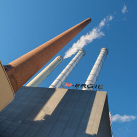 Das Bild zeigt ein Industriegebäude mit vier hohen Schornsteinen, die in den Himmel ragen. Die Schornsteine sind unterschiedlich gefärbt: rotbraun, weiß und zweimal silbern. Im Hintergrund ist ein klarer blauer Himmel mit wenigen Wolken zu sehen.