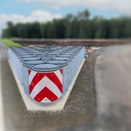 Das Bild zeigt eine Reihe von Anpralldämpfern am Ende einer Straße, die in dreieckiger Formation angeordnet sind. Die Frontflächen der Elemente tragen rot-weiße Pfeilmarkierungen. Im Hintergrund sind Bäume und eine angrenzende Straße zu erkennen.