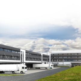 Das Bild zeigt einen großen Industrie- oder Gewerbebau mit mehreren Laderampen, an denen weiße Lastwagen geparkt sind. Das Gebäude hat mehrere Etagen mit vielen Fenstern. Im Vordergrund befindet sich eine Grasfläche, auf der zwei Personen spazieren gehen.