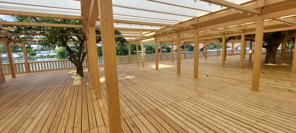 Das Bild zeigt eine große Holzterrasse mit mehreren Holzbalken und Pfosten, die ein durchsichtiges Dach tragen. In die Terrasse sind mehrere Bäume integriert, deren Stämme durch Aussparungen im Boden führen. Im Hintergrund ist Wasser zu sehen sowie Gebäude auf der gegenüberliegenden Uferseite.