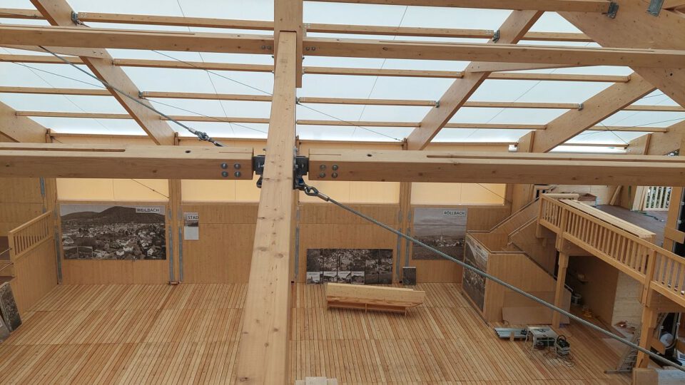 Das Bild zeigt das Innere eines Holzgebäudes mit hoher Decke, die von großen Holzbalken getragen wird. Der Boden besteht aus Holzplanken, an den Wänden hängen historische Schwarz-Weiß-Fotografien. Eine Treppe führt zu einer oberen Ebene mit Geländer, und das lichtdurchlässige Dach lässt Tageslicht in den Raum.