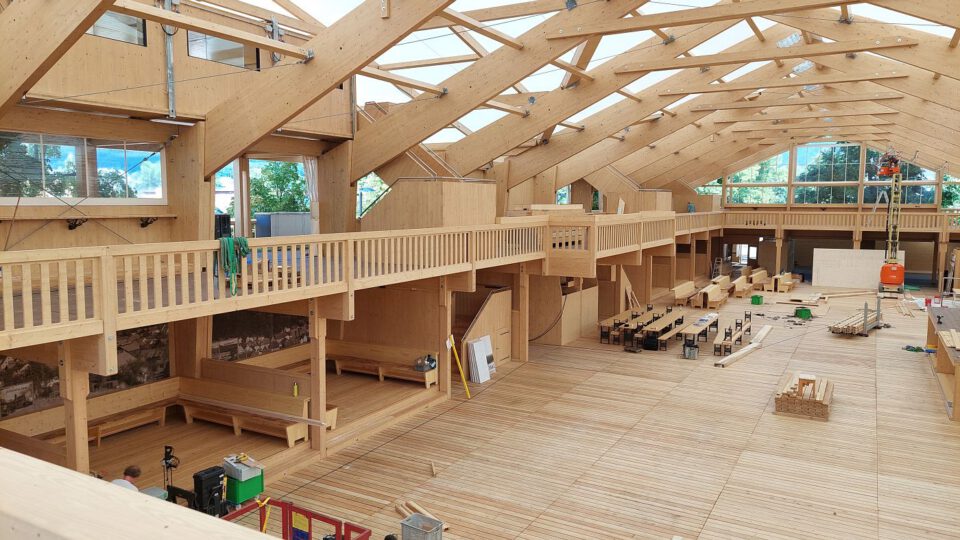 Das Bild zeigt das Innere eines großen Holzgebäudes im Bau mit sichtbaren Holzbalken und einer hohen Decke mit Oberlichtern. Eine umlaufende Galerie mit Geländer ist zu sehen, ebenso wie Werkbänke und Werkzeuge im Raum. Große Fenster auf beiden Seiten lassen viel Tageslicht herein.