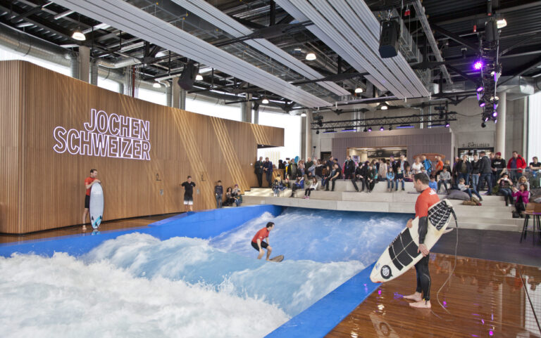Das Bild zeigt eine Indoor-Surfanlage mit einem Wellenbecken, in dem eine Person auf einer künstlichen Welle surft. Eine weitere Person steht am Beckenrand mit einem Surfbrett in der Hand. Im Hintergrund beobachten Zuschauer das Geschehen, und an der Holzwand ist groß der Name „Jochen Schweizer“ zu sehen.