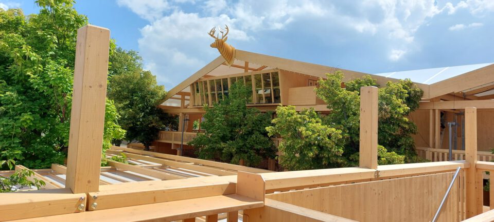 Das Bild zeigt ein mehrstöckiges Holzgebäude mit großen Fenstern und einer auffälligen Hirschskulptur auf dem Dach. Umgeben ist das Gebäude von Bäumen unter einem teils bewölkten Himmel.