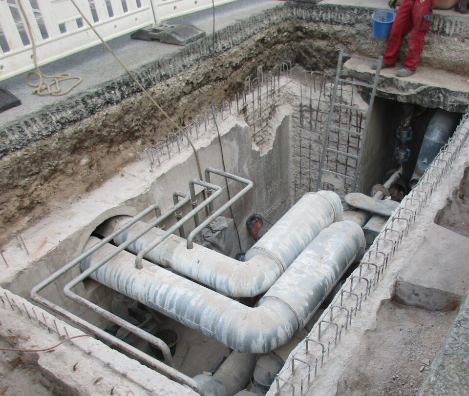 Das Bild zeigt eine Baustelle mit einem offenen Graben, in dem große Metallrohre und Bewehrungsstahl verlegt sind. Ein Arbeiter in roter Kleidung steht am Rand des Grabens, daneben befindet sich ein blauer Eimer. Die Szene gehört vermutlich zu einem Infrastrukturprojekt im Bereich Versorgung oder Entwässerung.