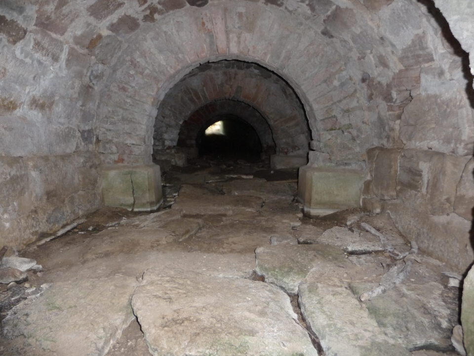 Das Bild zeigt einen alten Steintunnel mit gewölbter Decke und unebenem Boden aus großen, unregelmäßigen Steinplatten. Wände und Decke bestehen aus grob behauenen Steinen, was dem Tunnel ein rustikales Aussehen verleiht. In der Ferne sind zwei weitere Rundbögen zu sehen, durch die Licht einfällt.