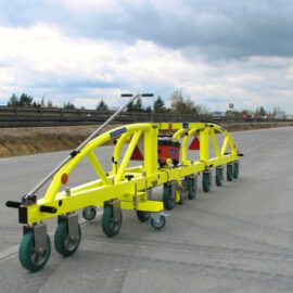 Das Bild zeigt ein gelbes, mehrrädriges Bau- oder Wartungsgerät auf einer asphaltierten Straße. Das Gerät besitzt mehrere grüne Räder und scheint für Arbeiten an der Straße eingesetzt zu werden.
