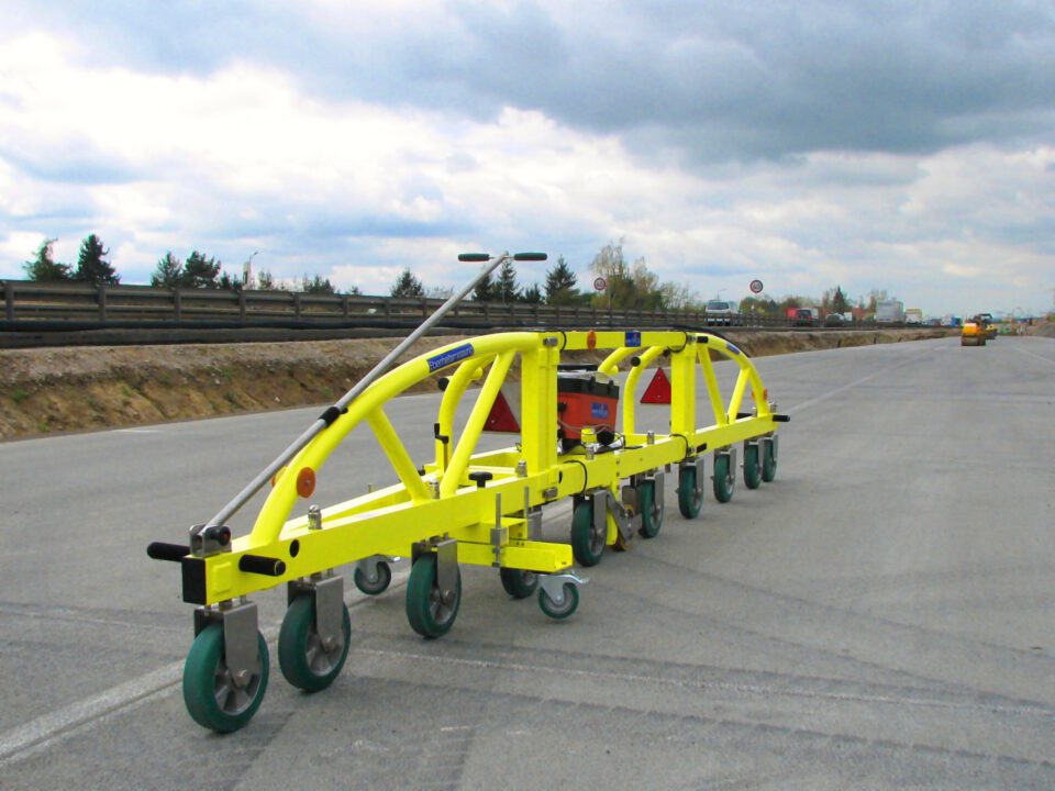 Das Bild zeigt ein gelbes, mehrrädriges Bau- oder Wartungsgerät auf einer asphaltierten Straße. Das Gerät besitzt mehrere grüne Räder und scheint für Arbeiten an der Straße eingesetzt zu werden.