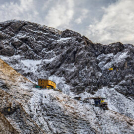 Das Bild zeigt eine verschneite Berglandschaft mit felsigen Hängen und einem Gebäude, das wie eine Berghütte aussieht. Ein gelber Hubschrauber fliegt in der Nähe des Hangs. Der Himmel ist teilweise bewölkt.