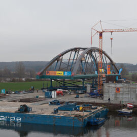 Das Bild zeigt eine Baustelle mit einer im Bau befindlichen Brücke über einem Gewässer. Auf der rechten Seite steht ein großer Kran, und auf dem Gelände sind verschiedene Baumaterialien und Geräte zu sehen. Im Vordergrund schwimmt ein blaues Bauschiff im Wasser, im Hintergrund sind Wiesen und Bäume unter einem bewölkten Himmel.
