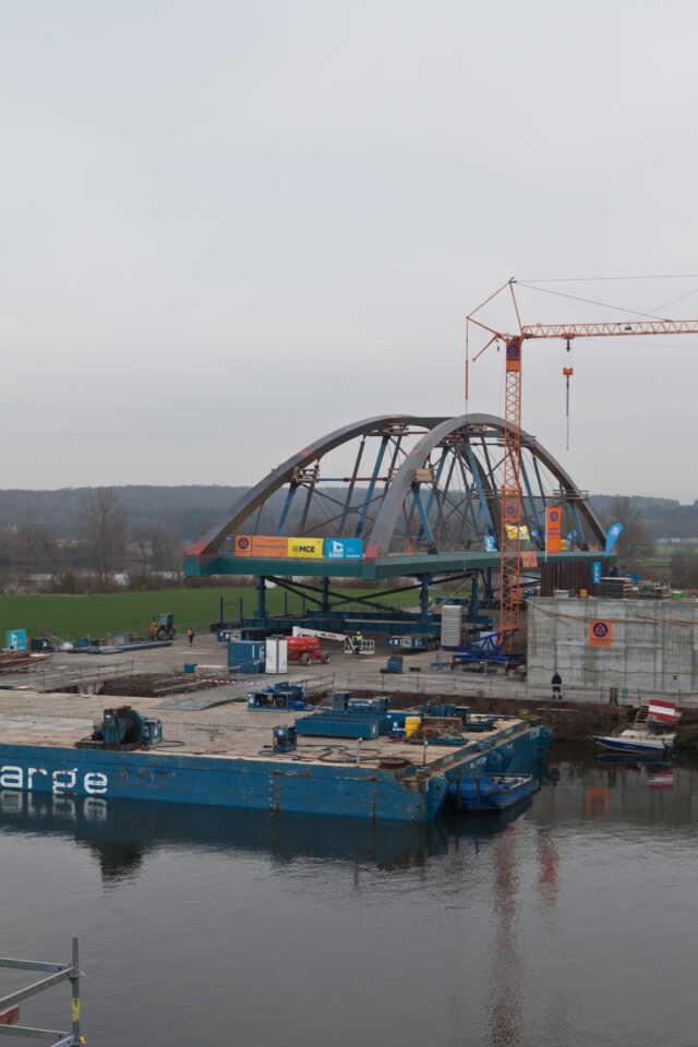 Das Bild zeigt eine Baustelle mit einer im Bau befindlichen Brücke über einem Gewässer. Auf der rechten Seite steht ein großer Kran, und auf dem Gelände sind verschiedene Baumaterialien und Geräte zu sehen. Im Vordergrund schwimmt ein blaues Bauschiff im Wasser, im Hintergrund sind Wiesen und Bäume unter einem bewölkten Himmel.