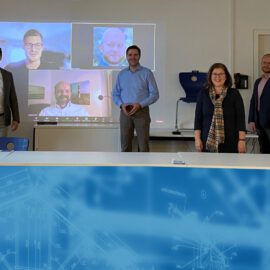 Das Bild zeigt eine Gruppe von neun Personen, die um einen Konferenztisch in einem Besprechungsraum stehen. Auf einem Bildschirm im Hintergrund sind vier weitere Teilnehmende virtuell zugeschaltet. Auf dem Tisch liegen Laptops, Dokumente, Wasserflaschen und ein technischer Plan oder eine Bauzeichnung.