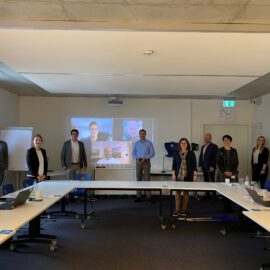 Das Bild zeigt einen Konferenzraum mit mehreren Personen, die um einen U-förmigen Tisch stehen. Neun Personen sind im Raum anwesend, vier weitere sind auf einem Bildschirm virtuell zugeschaltet. Der Raum verfügt über große Fenster, Whiteboards und verschiedene Büromaterialien auf den Tischen.