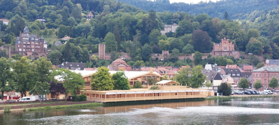 Das Bild zeigt eine malerische Stadtansicht an einem Fluss, umgeben von grünen Hügeln. Zu sehen sind Wohnhäuser und größere, teils historische Gebäude, eingebettet in dichte Vegetation. Im Vordergrund steht ein großes Holzgebäude am Flussufer, vermutlich ein Restaurant oder Veranstaltungsort mit Außenbereichen.