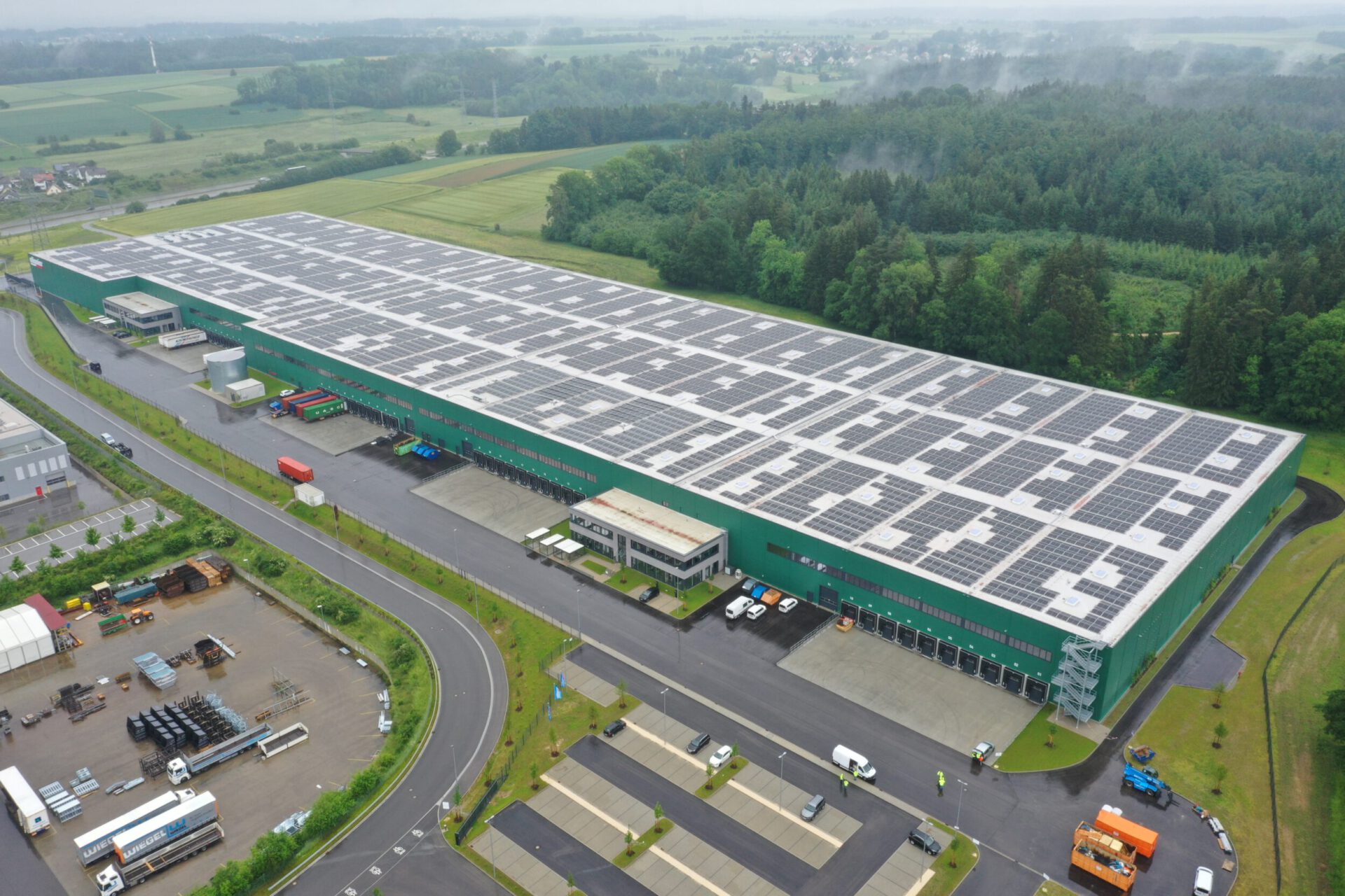 Das Bild zeigt ein großes Industriegebäude mit grüner Fassade und einer Dachfläche, die vollständig mit Solarpaneelen bedeckt ist. Rund um das Gebäude befinden sich Grünflächen, Bäume sowie geparkte Fahrzeuge und Lastwagen. In der Umgebung sind weitere kleinere Gebäude, Straßen und Parkflächen zu sehen, eingebettet in eine ländliche Landschaft mit Feldern und Wald.