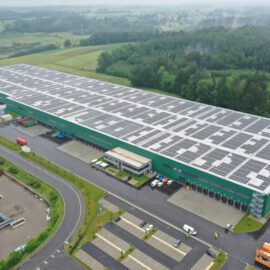 Das Bild zeigt ein großes Industriegebäude mit grüner Fassade und einer Dachfläche, die vollständig mit Solarpaneelen bedeckt ist. Rund um das Gebäude befinden sich Grünflächen, Bäume sowie geparkte Fahrzeuge und Lastwagen. In der Umgebung sind weitere kleinere Gebäude, Straßen und Parkflächen zu sehen, eingebettet in eine ländliche Landschaft mit Feldern und Wald.