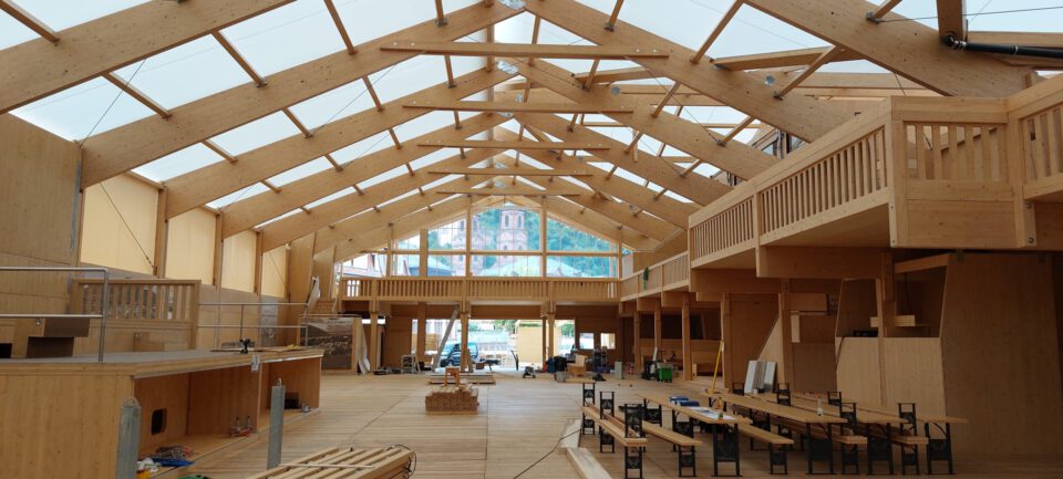 Das Bild zeigt das Innere eines großen Holzgebäudes mit hohen Decken und sichtbaren Holzbalken. Auf mehreren Ebenen befinden sich Holzgeländer und Balkone, während im Erdgeschoss Tische, Stühle und Baumaterialien verteilt sind. Durch große Fenster im Hintergrund ist eine Außenanlage mit Bäumen und einem Gebäude zu sehen.