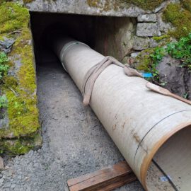 Das Bild zeigt ein großes zylindrisches Rohr, das in einen Tunneleingang aus Stein und Beton eingesetzt oder dort platziert wird. Um den Tunnel herum sind moosbewachsene Wände sowie Erde und Schotter zu sehen. Holzplanken und Werkzeuge in der Nähe des Rohrs deuten auf laufende Bau- oder Wartungsarbeiten hin.