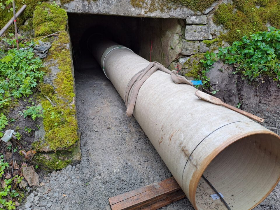 Das Bild zeigt ein großes zylindrisches Rohr, das in einen Tunneleingang aus Stein und Beton eingesetzt oder dort platziert wird. Um den Tunnel herum sind moosbewachsene Wände sowie Erde und Schotter zu sehen. Holzplanken und Werkzeuge in der Nähe des Rohrs deuten auf laufende Bau- oder Wartungsarbeiten hin.