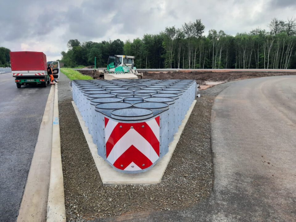 Das Bild zeigt eine Straßenbaustelle mit mehreren Metallfässern in dreieckiger Anordnung am Ende einer Betonleitwand. Die Fässer tragen rot-weiße Chevron-Reflektoren. Links im Bild steht ein Lkw mit zwei Arbeitern, im Hintergrund sind Baumaschinen und eine vorbereitete Arbeitsfläche zu sehen. Der Himmel ist bewölkt, im Hintergrund befinden sich Bäume.