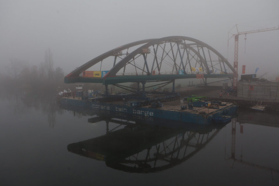 Das Bild zeigt eine große, bogenförmige Brückenkonstruktion, die auf einem Lastschiff über ein Gewässer transportiert wird. Nebel liegt über der Szene und sorgt für eine eingeschränkte Sicht, während am Ufer Baugeräte und ein Kran zu sehen sind.