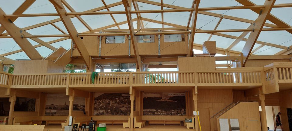 Das Bild zeigt das Innere eines Holzgebäudes mit offenem Dach und sichtbaren Holzbalken. Auf einer zweiten Ebene befinden sich Räume mit Holzgeländern, während im unteren Bereich Bänke sowie großformatige Fotos oder Wandbilder mit historischen Motiven zu sehen sind. Stühle und weitere Gegenstände sind im Raum verteilt.