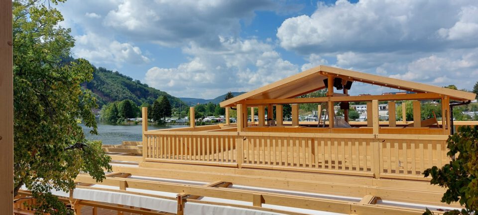 Das Bild zeigt eine hölzerne Überdachung mit offenen Seiten, die sich in der Nähe eines Gewässers befindet. Die Konstruktion wirkt wie ein überdachter Sitz- oder Aussichtspunkt im Freien, möglicherweise für Veranstaltungen. Umgeben ist sie von Bäumen, im Hintergrund sind grüne Hügel unter einem teils bewölkten Himmel zu sehen.