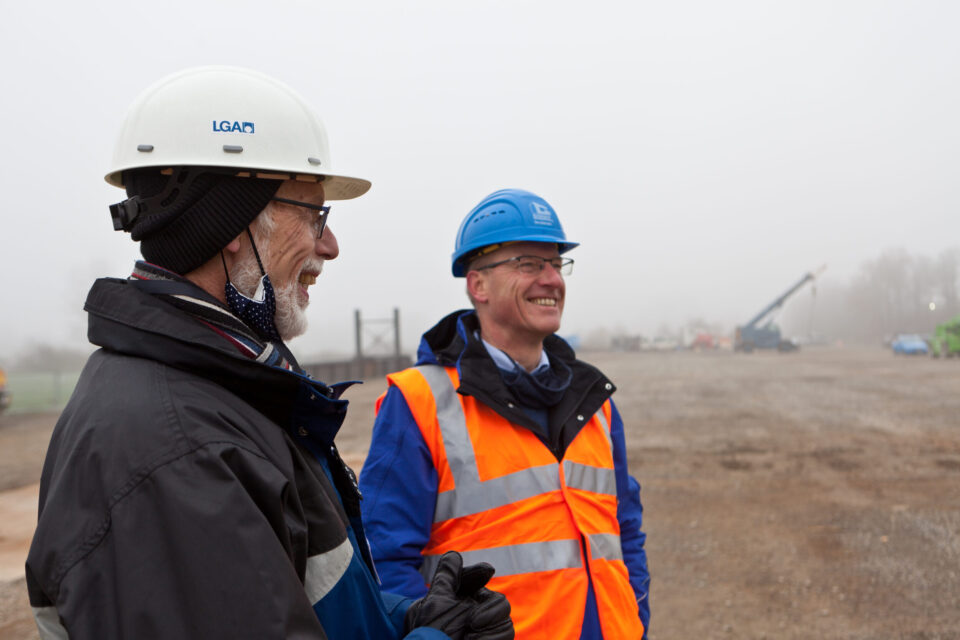 Das Bild zeigt zwei Personen mit Sicherheitshelmen und Warnkleidung auf einer Baustelle. Im Hintergrund sind Baumaschinen und Fahrzeuge zu sehen, die Szene ist in Nebel gehüllt.