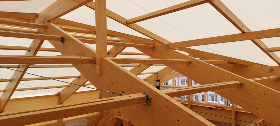 Das Bild zeigt eine detaillierte Ansicht einer Holzdachkonstruktion mit zahlreichen Balken und Streben, die durch Metallverbindungen und Schrauben gesichert sind. Die Holzbalken sind in einem komplexen Muster angeordnet, um Stabilität und Tragkraft zu gewährleisten. Zusätzlich verlaufen Kabel oder Drähte durch die Konstruktion, vermutlich zur zusätzlichen Sicherung.