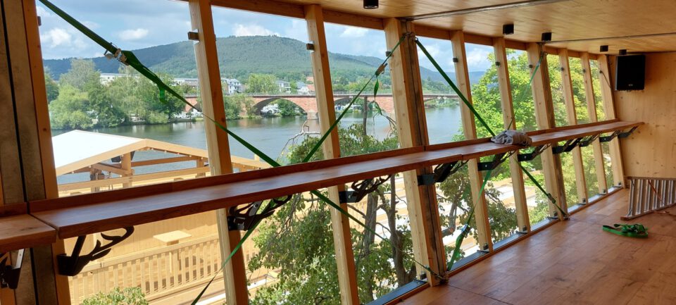 Das Bild zeigt das Innere eines Holzgebäudes mit großen Fenstern, die den Blick auf eine Flusslandschaft freigeben. Die Fenster sind mit grünen Gurten an den Holzbalken befestigt. Im Hintergrund sind ein Fluss, eine Brücke sowie Bäume und Hügel zu sehen.