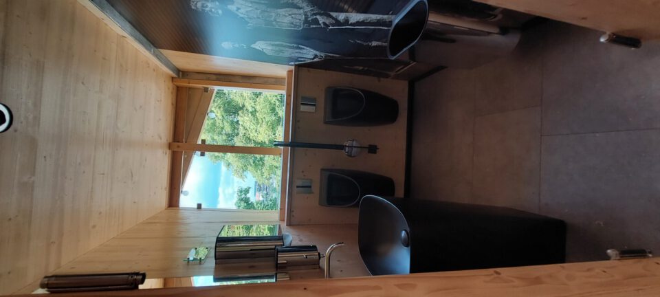 Das Bild zeigt das Innere einer modernen Toilettenanlage mit Holzverkleidung an Wänden und Decke. An der Wand befinden sich zwei schwarze Urinale sowie ein schwarzes Waschbecken mit Wasserhahn und Spiegel darüber. Ein offenes Fenster gibt den Blick auf grüne Außenanlagen frei, und an einer Wand ist ein künstlerisches Wandbild mit stehenden Personen zu sehen.