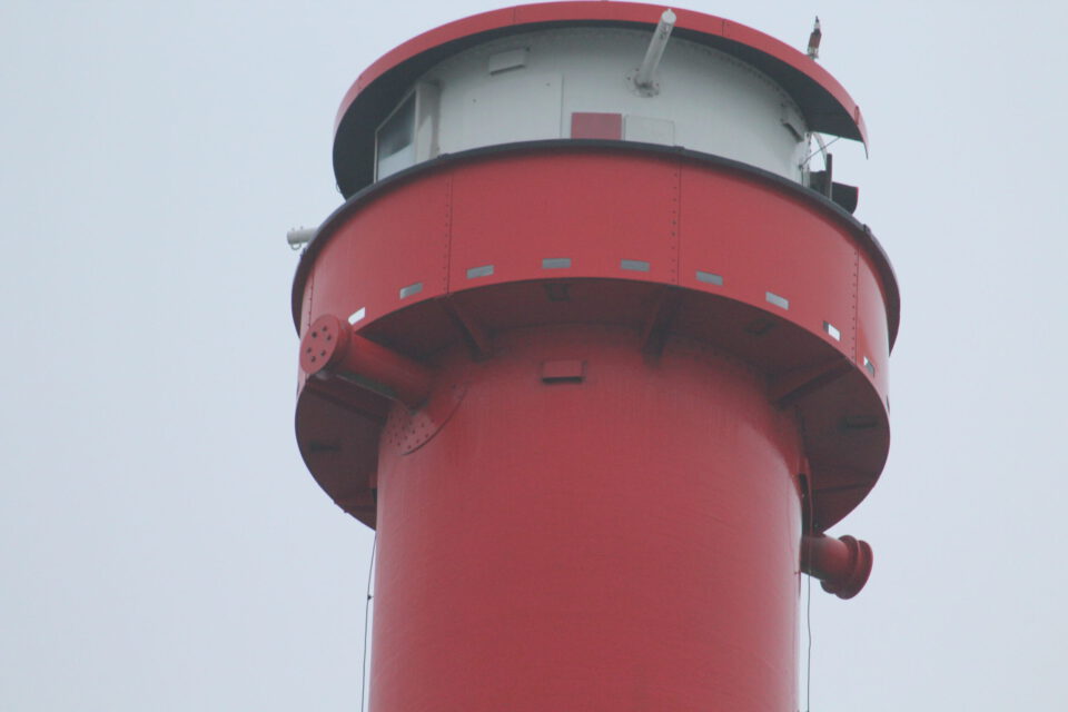 Das Bild zeigt den oberen Teil eines roten Leuchtturms mit einem runden Aussichtsbalkon unterhalb der Spitze. Der Balkon verfügt über kleine rechteckige Fenster und ist mit einem roten Dach bedeckt. Außen am Turm sind verschiedene bauliche Elemente wie Rohre und Träger sichtbar.