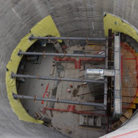 Das Bild zeigt eine Baustelle im Inneren eines großen zylindrischen Bauwerks mit Betonwänden. Quer über den Zylinder verlaufen mehrere horizontale Stahlträger, dazu sind rote Gerüste, Leitern und gelbe Schutzabdeckungen an den Wänden zu sehen. Am Boden befinden sich Bauarbeiter sowie verschiedene Baumaterialien und Geräte.