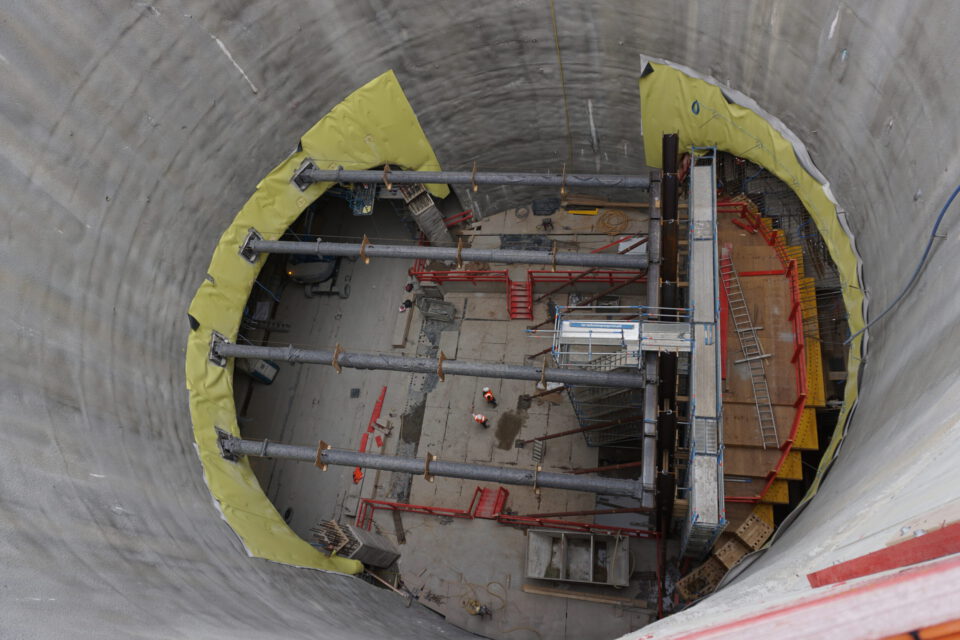 Das Bild zeigt eine Baustelle im Inneren eines großen zylindrischen Bauwerks mit Betonwänden. Quer über den Zylinder verlaufen mehrere horizontale Stahlträger, dazu sind rote Gerüste, Leitern und gelbe Schutzabdeckungen an den Wänden zu sehen. Am Boden befinden sich Bauarbeiter sowie verschiedene Baumaterialien und Geräte.