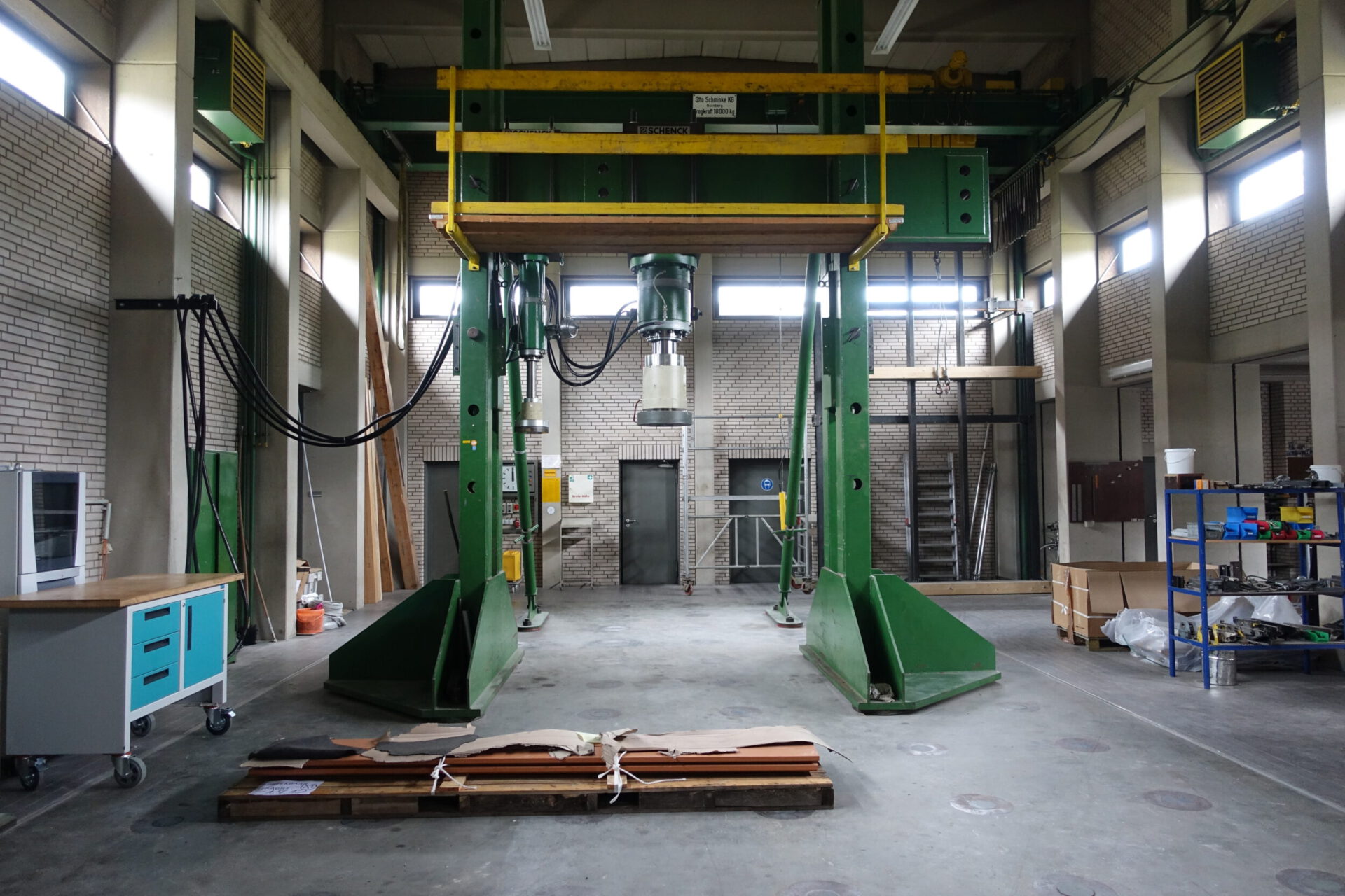 Das Bild zeigt eine industrielle Werkhalle mit einer großen grün-gelben Maschine in der Mitte, vermutlich eine hydraulische Presse. Umgeben ist sie von Kabeln, einem blauen Werkstattwagen mit Schubladen, einem Metallregal mit Werkzeugen und gestapeltem Material. Im Hintergrund sind Backsteinwände, Fenster und Türen zu weiteren Bereichen der Halle zu sehen.