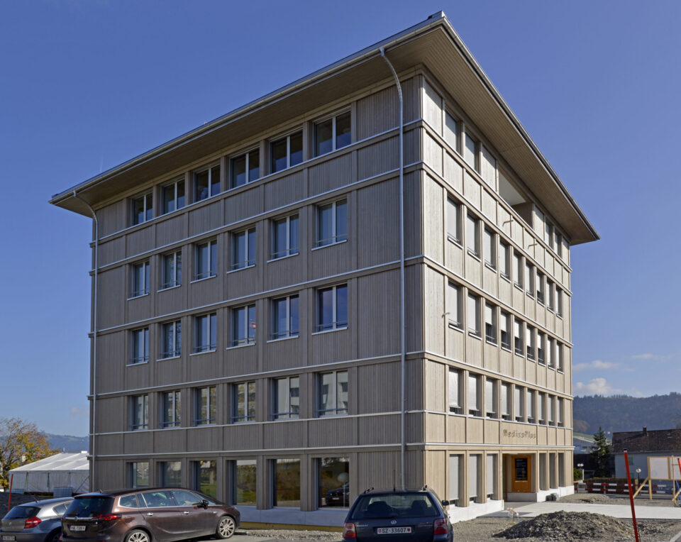 Das Bild zeigt ein mehrstöckiges Gebäude mit moderner Architektur, großen Fenstern und einer Holzfassade. Vor dem Gebäude sind mehrere Autos geparkt, im Hintergrund ist etwas Grün zu sehen. Der Himmel ist klar und blau, was auf Tageslicht und gutes Wetter hinweist.