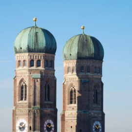 Das Bild zeigt die beiden Türme der Frauenkirche in München mit ihren markanten grünen Kupferhauben und gut sichtbaren Uhren. Die Architektur ist klar erkennbar vor einem strahlend blauen Himmel.
