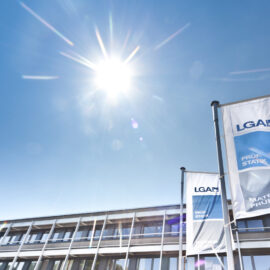 Das Bild zeigt ein modernes Gebäude mit großen Fenstern unter einem klaren blauen Himmel bei Sonnenschein. Vor dem Gebäude stehen zwei hohe Fahnenmasten mit jeweils einer Fahne.