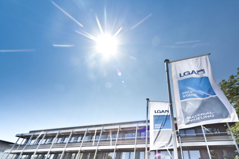 Das Bild zeigt ein modernes Gebäude mit großen Fenstern unter einem klaren blauen Himmel bei Sonnenschein. Vor dem Gebäude stehen zwei hohe Fahnenmasten mit jeweils einer Fahne.