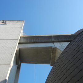 Das Bild zeigt eine moderne Architekturaufnahme aus der Froschperspektive mit Beton- und Metallstrukturen. Links ist eine hohe Betonwand mit zwei Metallbefestigungen zu sehen, in der Mitte ein waagerechter Betonträger, der zu einem weiteren Bauteil führt. Rechts befindet sich eine gewölbte Fläche mit dunkler Verkleidung vor blauem Himmel.