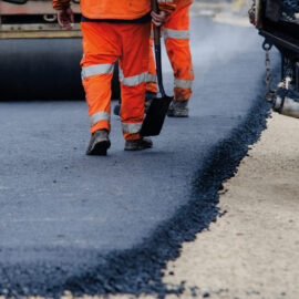 Das Bild zeigt zwei Arbeiter in orangefarbener Warnkleidung auf einer Straßenbaustelle. Einer der Arbeiter hält eine Schaufel, im Hintergrund steht eine Straßenwalze. Frisch aufgetragener Asphalt ist deutlich am Übergang zur helleren Tragschicht zu erkennen.
