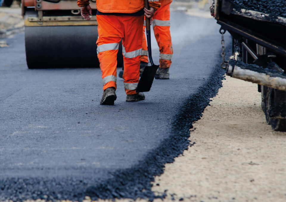 Das Bild zeigt zwei Arbeiter in orangefarbener Warnkleidung auf einer Straßenbaustelle. Einer der Arbeiter hält eine Schaufel, im Hintergrund steht eine Straßenwalze. Frisch aufgetragener Asphalt ist deutlich am Übergang zur helleren Tragschicht zu erkennen.