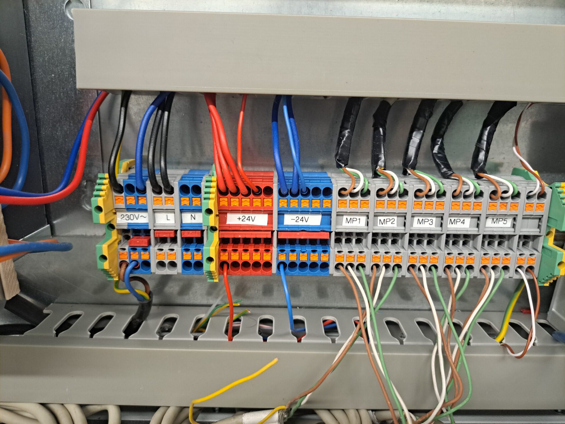 Das Bild zeigt eine elektrische Anschlussleiste mit mehreren farbigen Kabeln, die daran angeschlossen sind. Die Klemmen sind nebeneinander angeordnet und tragen unterschiedliche Bezeichnungen. Die Kabel sind farblich codiert, unter anderem in Blau, Schwarz und Rot.