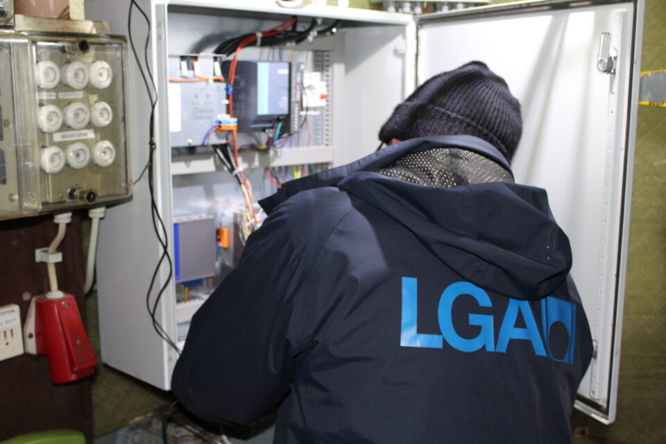 Eine Person arbeitet an einem geöffneten elektrischen Schaltschrank, der mit zahlreichen Kabeln, Platinen und elektronischen Bauteilen bestückt ist. Die Person trägt eine Jacke mit der Aufschrift LGA auf dem Rücken. Links im Bild ist ein altmodisches Telefonvermittlungsgerät zu sehen.