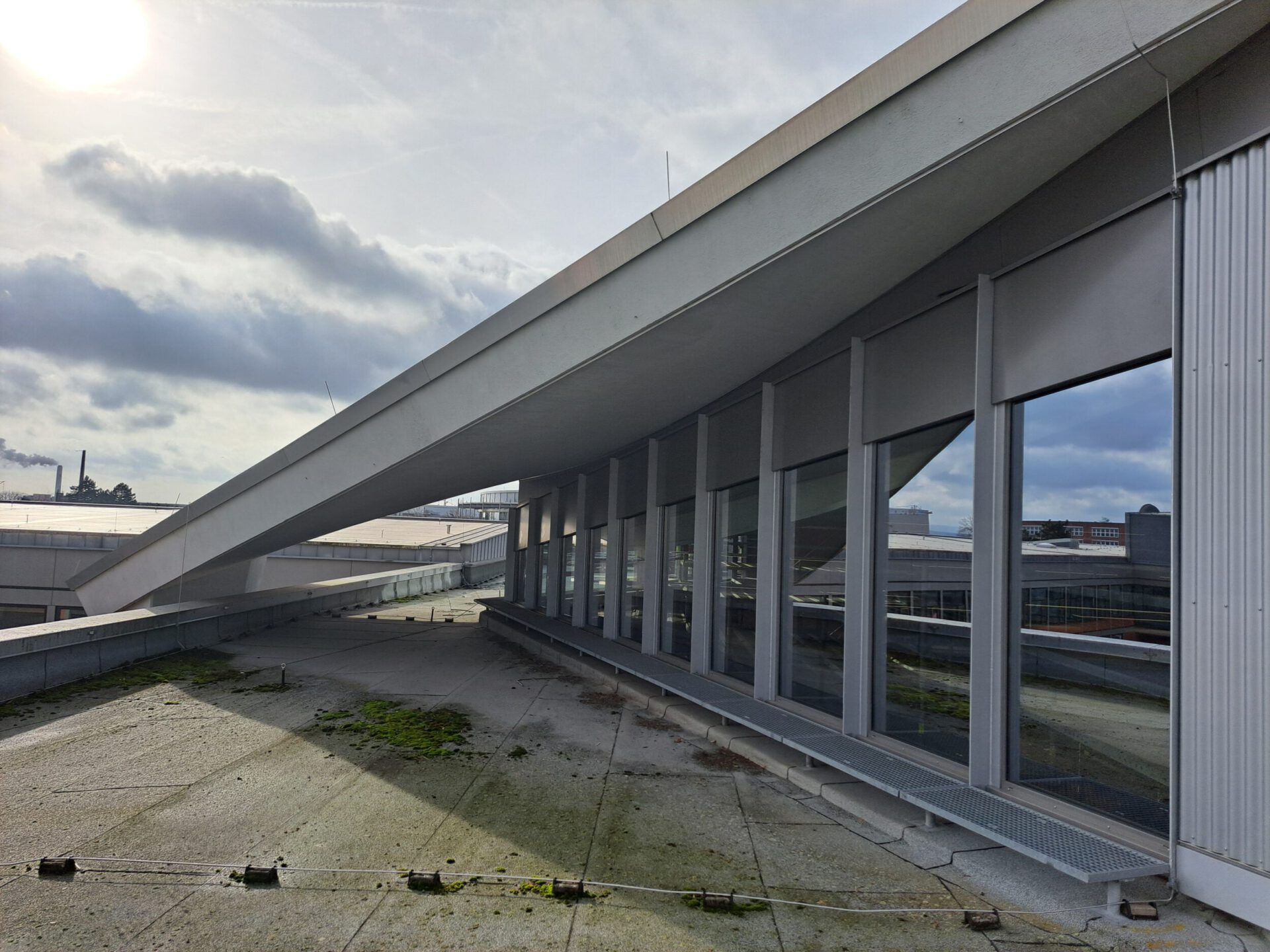 Das Bild zeigt eine Dachfläche mit einem schrägen Dachaufbau, der sich über eine Reihe großer Fenster erstreckt. Der Boden besteht aus Beton mit vereinzelten Moos- oder Pflanzenflecken. Im Hintergrund sind Industriegebäude und ein Schornstein unter einem teils bewölkten Himmel mit Sonnenschein zu sehen.