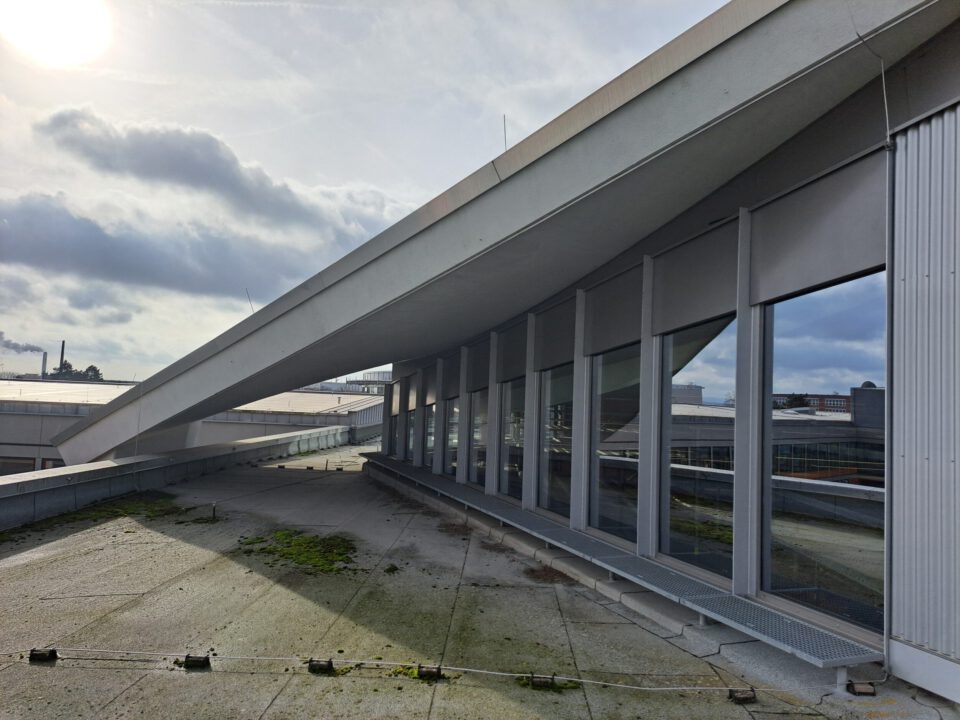 Das Bild zeigt eine Dachfläche mit einem schrägen Dachaufbau, der sich über eine Reihe großer Fenster erstreckt. Der Boden besteht aus Beton mit vereinzelten Moos- oder Pflanzenflecken. Im Hintergrund sind Industriegebäude und ein Schornstein unter einem teils bewölkten Himmel mit Sonnenschein zu sehen.