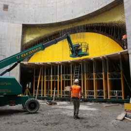 Das Bild zeigt eine Baustelle im Inneren eines großen Betonbauwerks. Im Vordergrund steht ein Arbeiter mit orangefarbener Warnweste und weißem Helm vor einem grünen Bau-Lift, dessen Plattform an einer gelben, gebogenen Schalung positioniert ist. Die Schalung wird von Gerüsten und Holzbalken gestützt, rundherum liegen verschiedene Baumaterialien und Geräte.