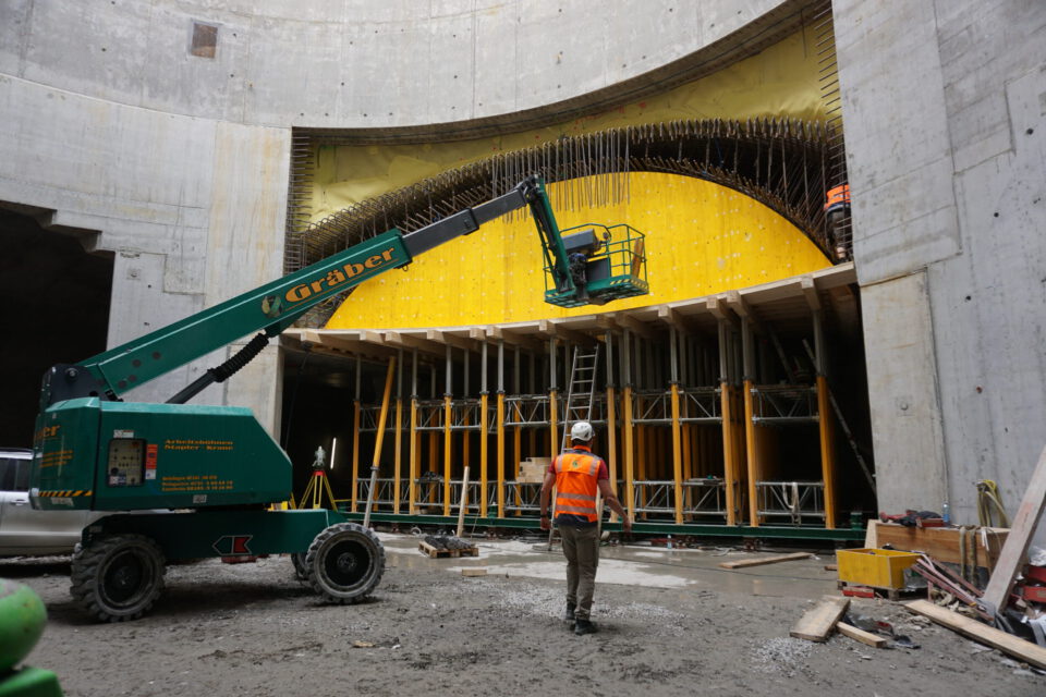 Das Bild zeigt eine Baustelle im Inneren eines großen Betonbauwerks. Im Vordergrund steht ein Arbeiter mit orangefarbener Warnweste und weißem Helm vor einem grünen Bau-Lift, dessen Plattform an einer gelben, gebogenen Schalung positioniert ist. Die Schalung wird von Gerüsten und Holzbalken gestützt, rundherum liegen verschiedene Baumaterialien und Geräte.