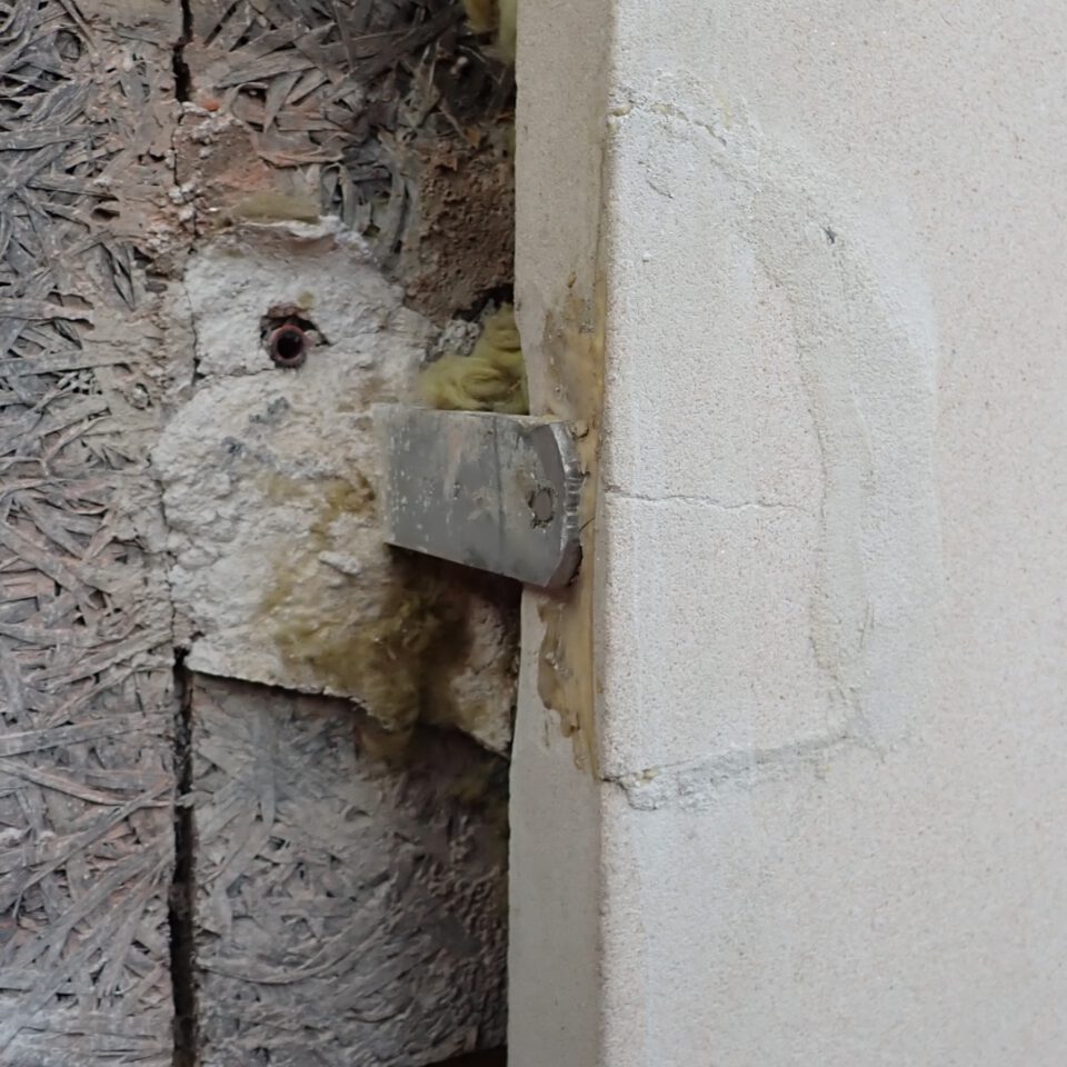 Das Bild zeigt eine Nahaufnahme einer Gebäudeecke mit sichtbaren Baumaterialien. Links ist eine offene Wandstelle mit Dämmmaterial und einer Holzwerkstoffplatte zu sehen, an der ein Metallwinkel befestigt ist. Rechts befindet sich eine verputzte Fläche mit Rissen und Spuren von Ausbesserungen rund um die Befestigung.