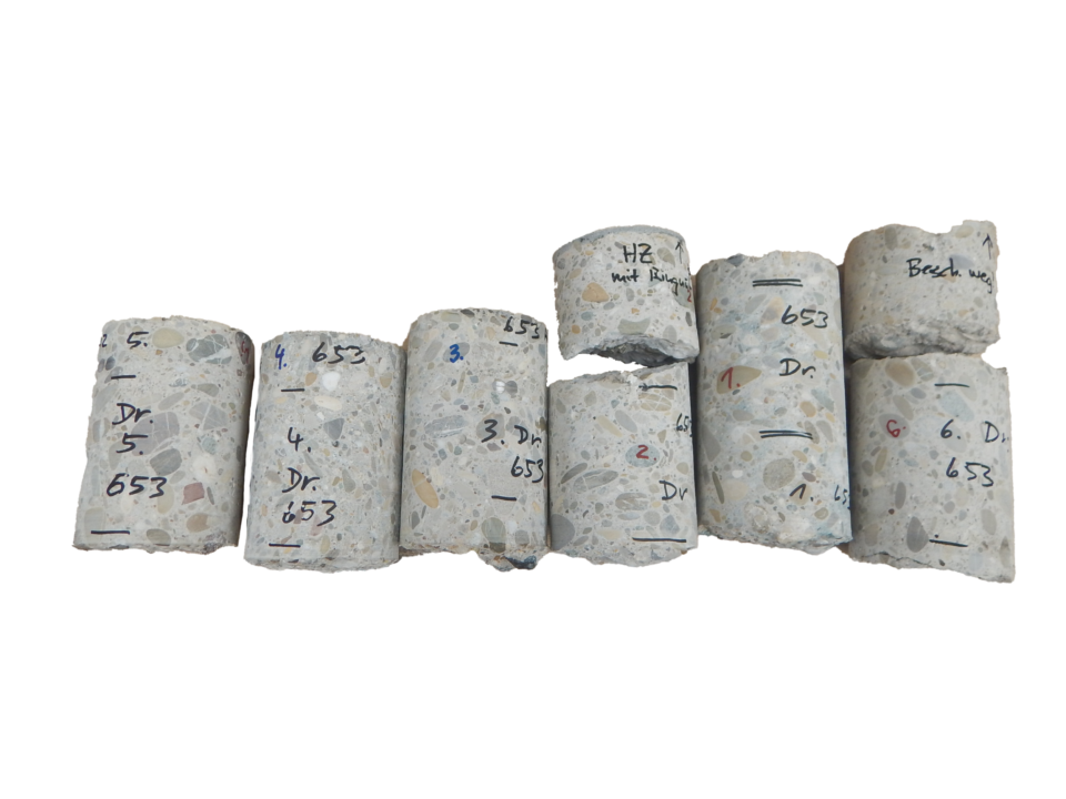 Das Bild zeigt acht zylindrische Betonproben, die in einer Reihe angeordnet sind. Einige der Proben weisen Risse oder Bruchstellen auf, was auf durchgeführte Belastungstests hindeutet. Auf den Proben sind handschriftliche Kennzeichnungen mit Zahlen und Buchstaben zu sehen.